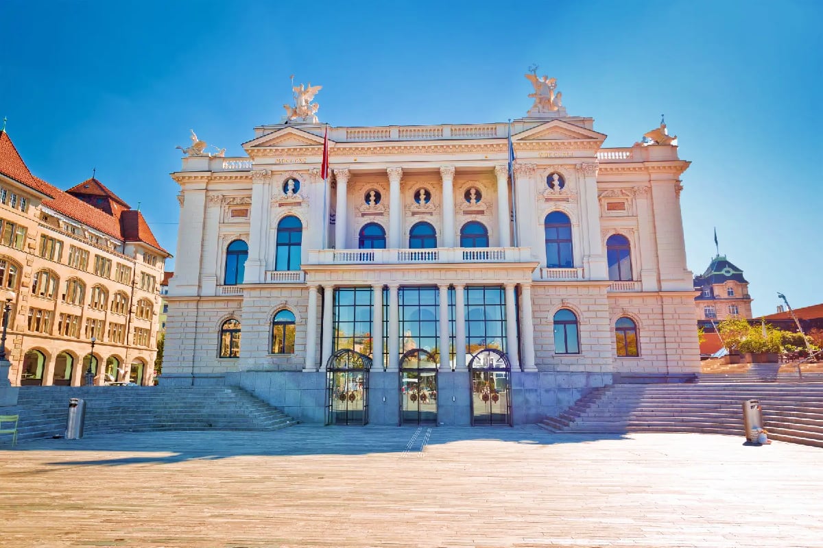 Grand neoclassical theater building with white columns and ornate sculptures, surrounded by historic architecture in a Europe