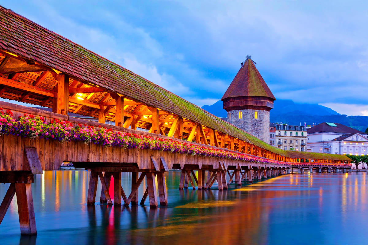 Chapel Bridge in Lucerne, Switzerland at dusk with wooden covered bridge, flower baskets, and tower reflected in calm water