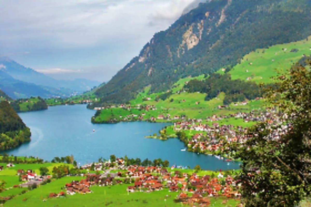 Alpine valley village with turquoise lake surrounded by green meadows, forested mountains, and traditional chalets with red r