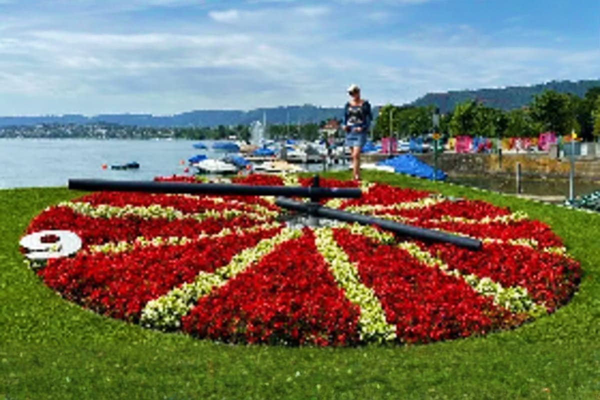 Large red and white flower clock display on lakeside green lawn with boats, mountains and city skyline in background