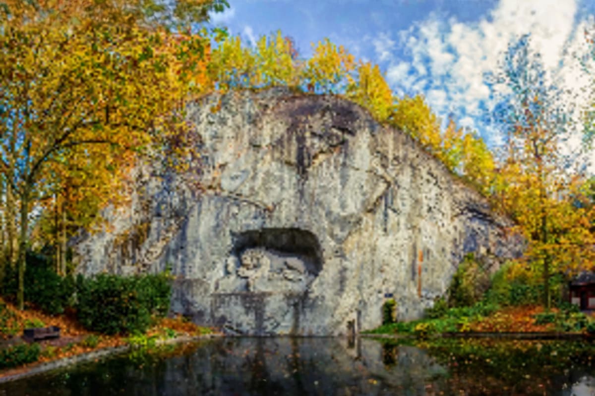 Giant stone head sculpture carved into cliff reflected in calm water, surrounded by autumn trees with yellow foliage