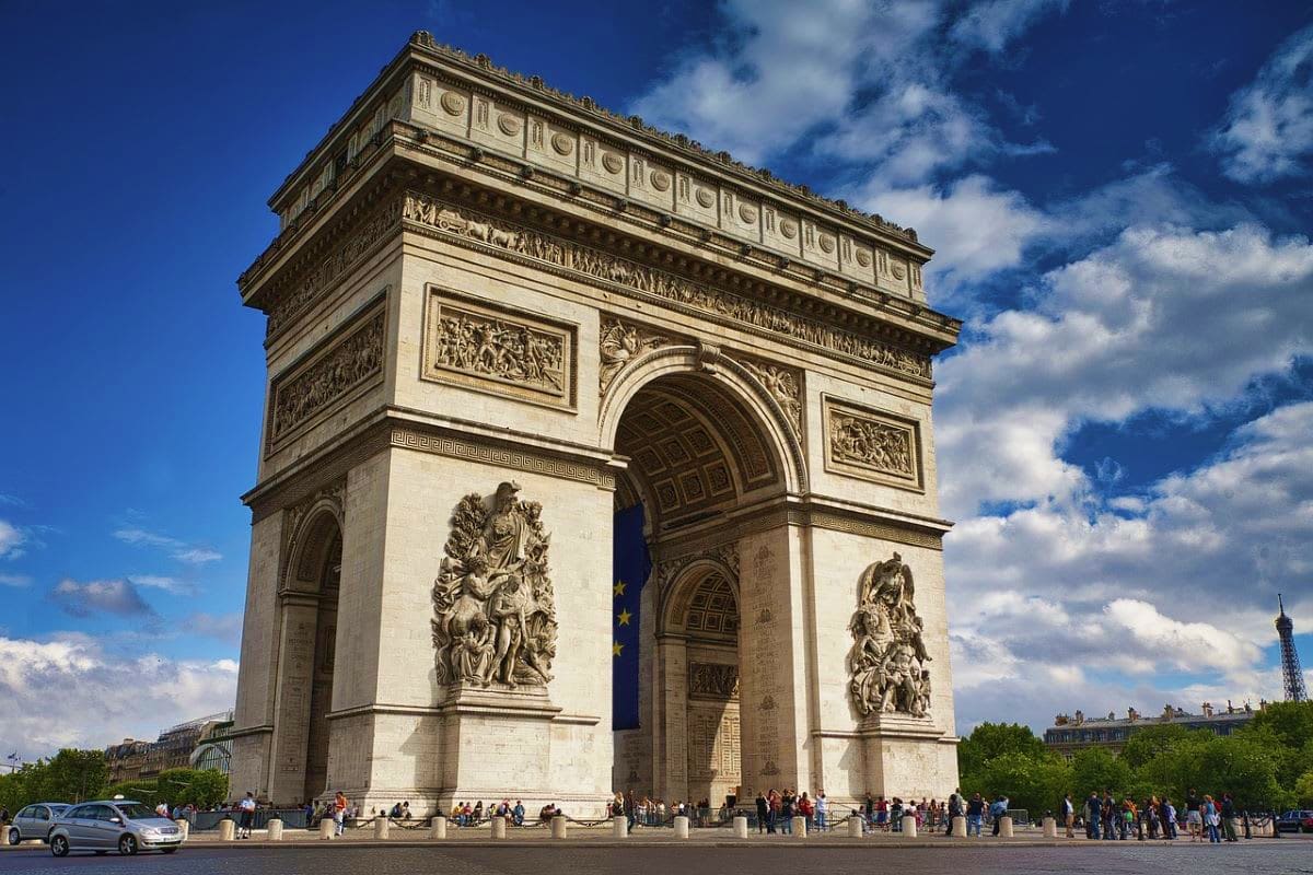 Arc de Triomphe monument in Paris with ornate stone facade, sculptural reliefs, and tourists gathered at its base under blue 