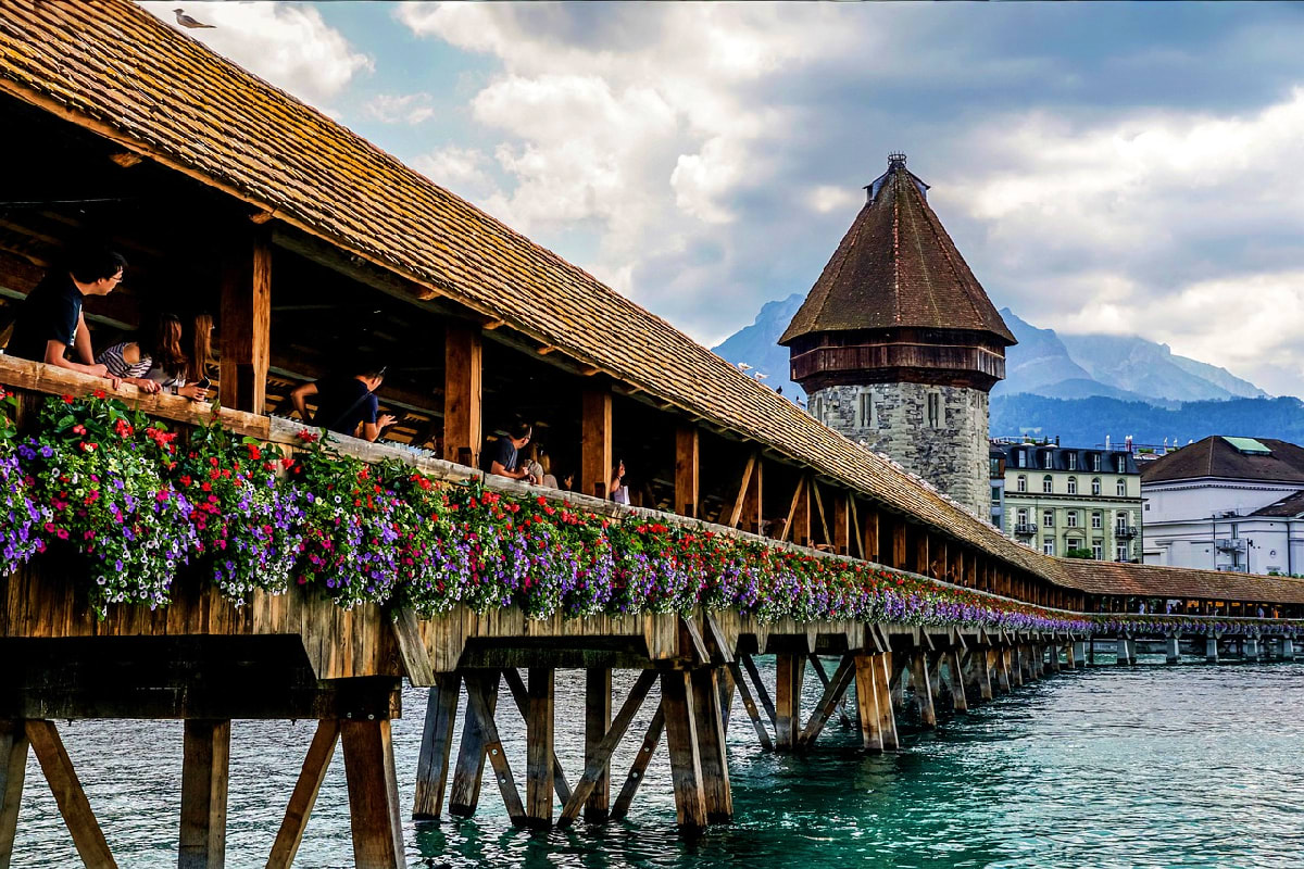 Historic wooden bridge with red and purple flower baskets spanning turquoise water, with conical tower and Alpine mountains i