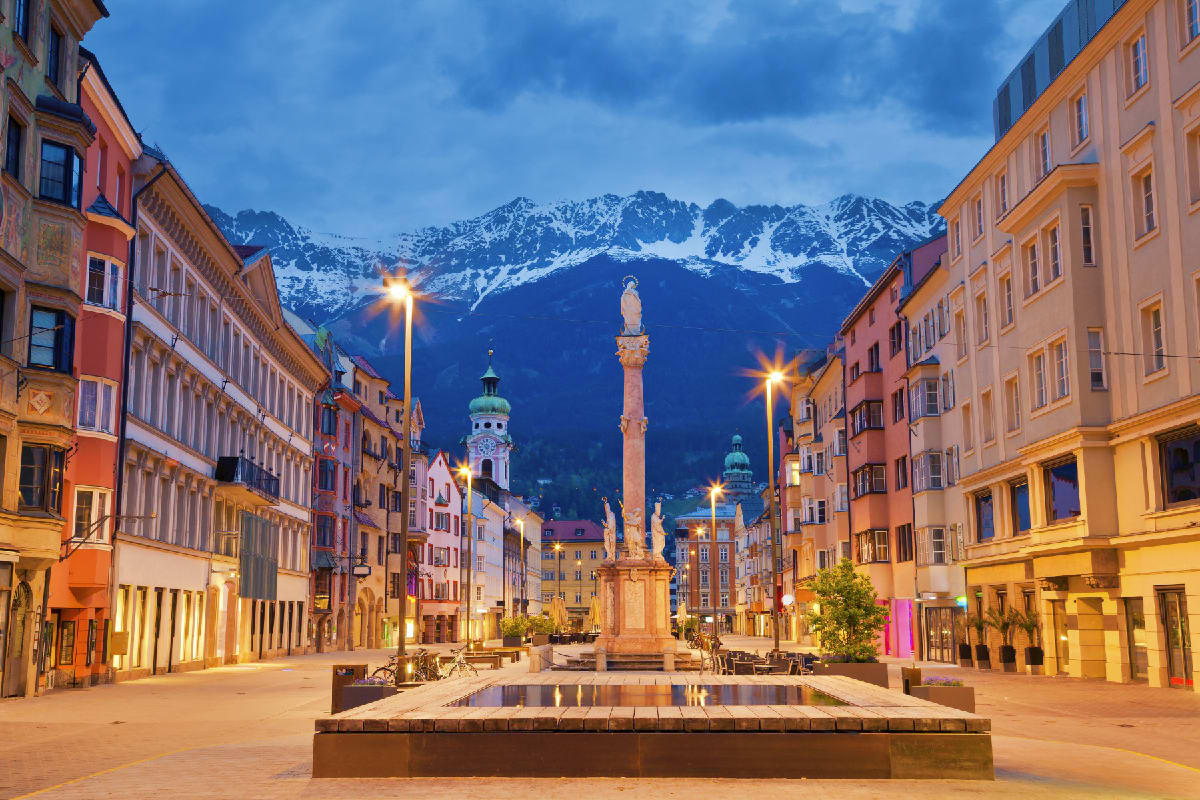 Best of Italy Switzerland and France Innsbruck city center at twilight with colorful buildings, illuminated street lamps, historic monument, and snow-capped Alpin