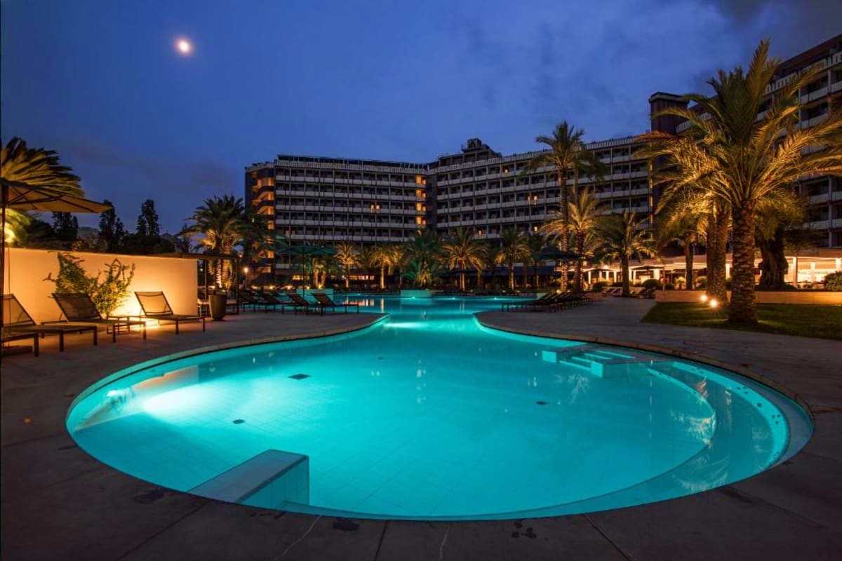 Illuminated resort pool at night with palm trees, lounge chairs, and multi-story hotel buildings in background under moonlit 