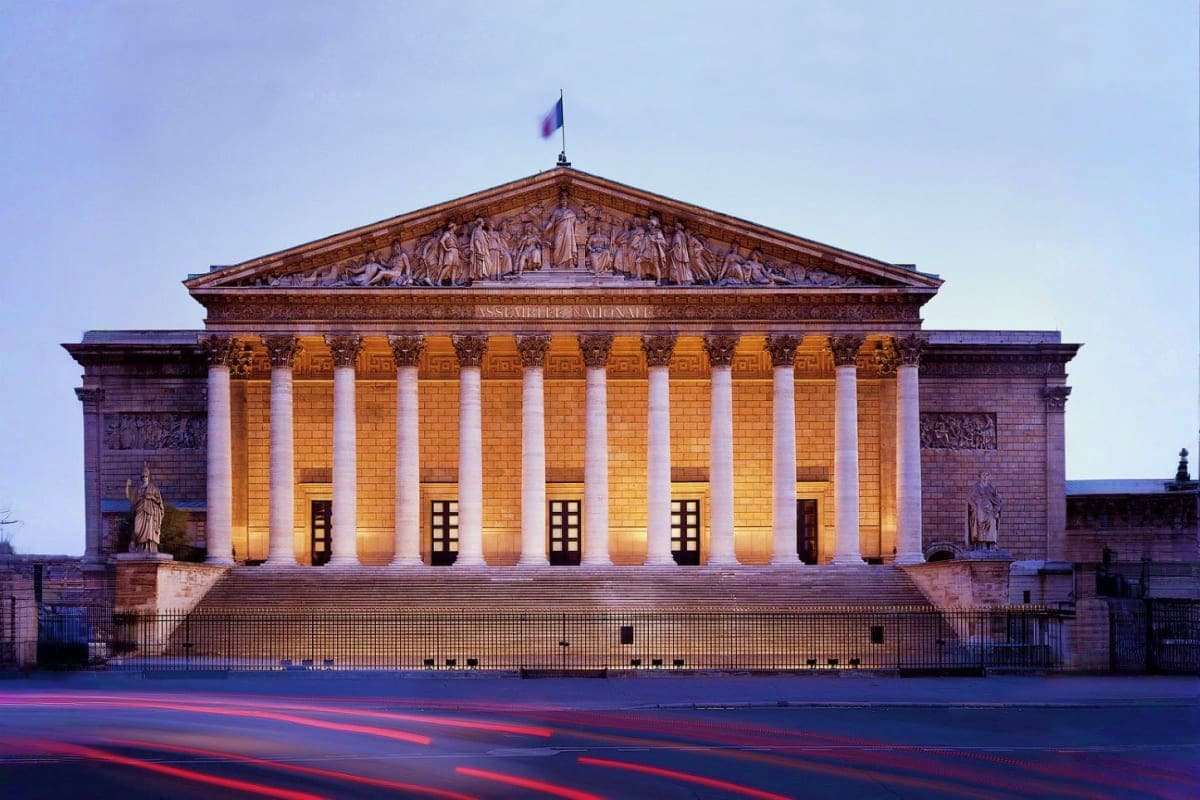 French National Assembly building with classical columns and pediment, illuminated at dusk with traffic light trails in foreg