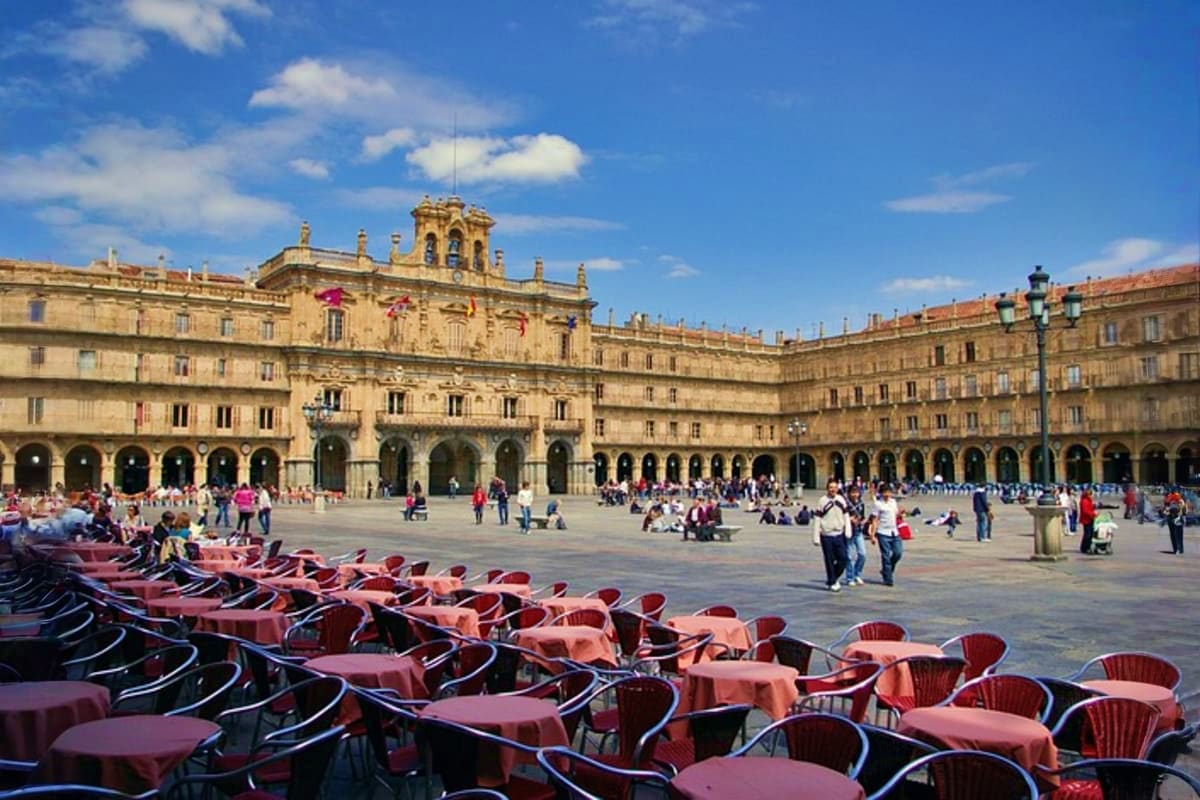 Historic plaza with golden Renaissance buildings, outdoor café seating, and tourists gathering under blue sky in Salamanca, S