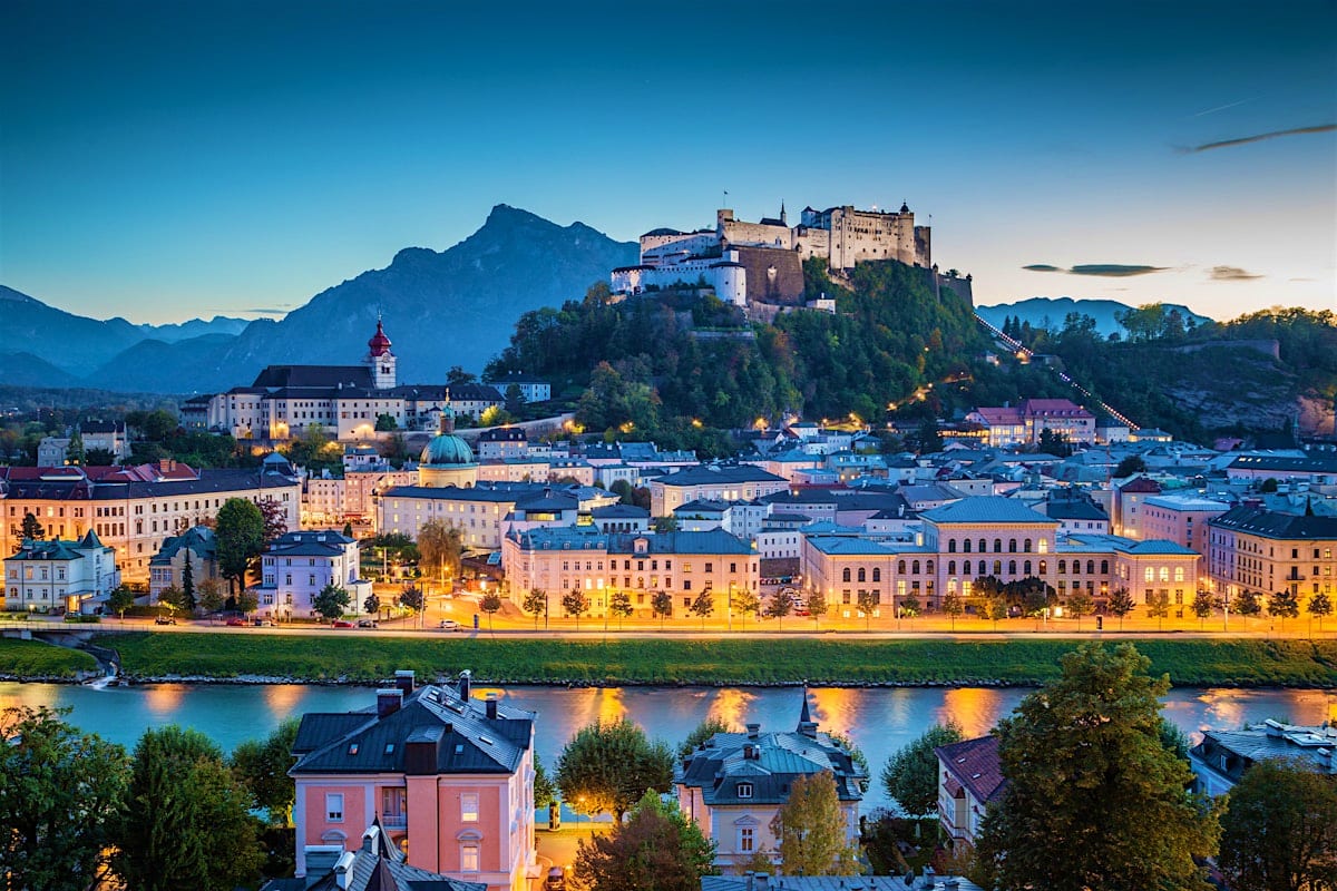 Italy Austria Journey Salzburg historic city skyline at dusk with Hohensalzburg Fortress on hilltop, illuminated buildings, and Salzach River in fo