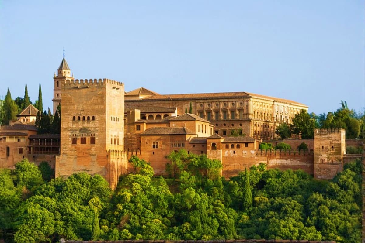 Alhambra palace in Granada, Spain, with golden stone fortress towers and walls surrounded by green trees under clear blue sky