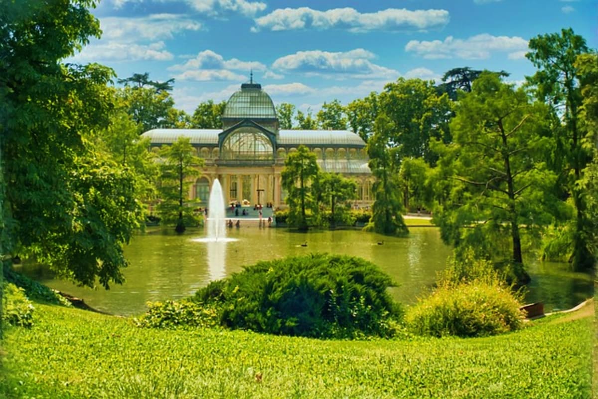 Crystal Palace in Retiro Park, Madrid, surrounded by lush green trees and a fountain lake on a sunny day