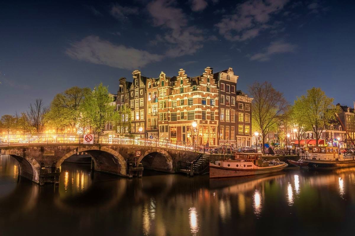 Historic Amsterdam canal with illuminated Dutch Renaissance buildings, arched stone bridge, and moored boats at dusk