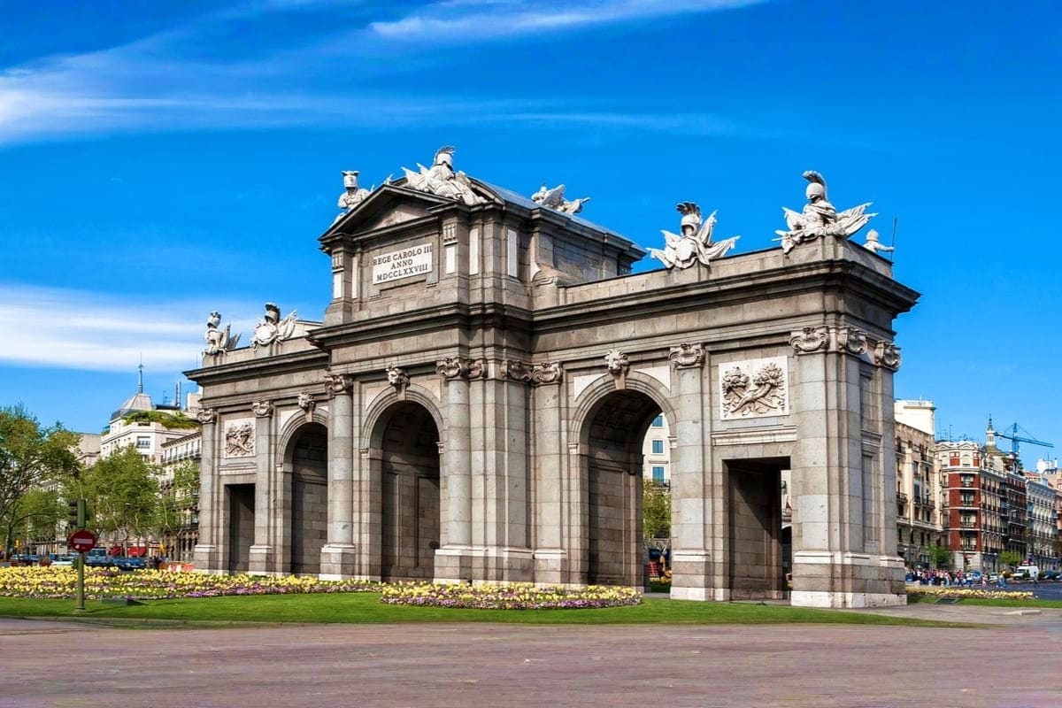 Puerta de Alcalá, a grand neoclassical stone gateway monument in Madrid, Spain, with blue sky and yellow flowers below.