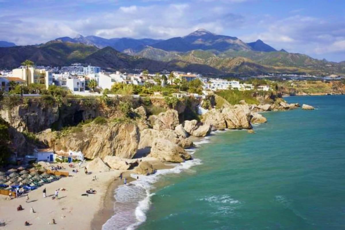 Nerja beach with white-washed buildings on clifftop, turquoise Mediterranean waters, rocky coastline and mountains in backgro