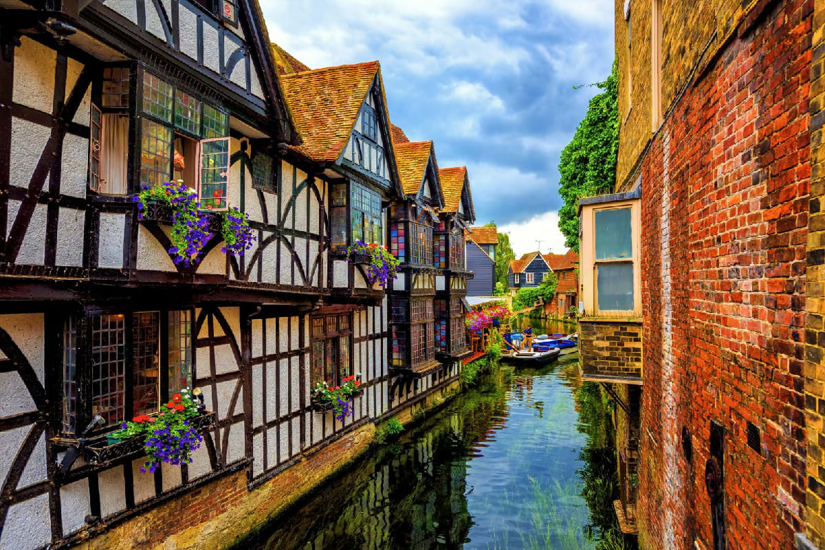 Historic Tudor buildings with purple flowers along a narrow canal in a charming English village