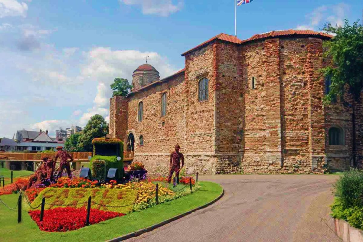 Historic Roman stone castle with red brick tower and flag, surrounded by colorful flower beds and manicured gardens in Colche