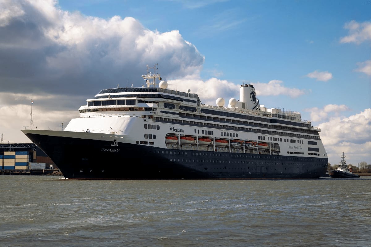 Large cruise ship Volendam docked at port with tugboat assistance, white and black hull against cloudy sky and calm water