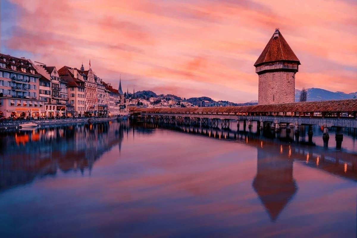 Lucerne waterfront at sunset with historic tower and wooden bridge reflected in calm lake water, Swiss Alps in background