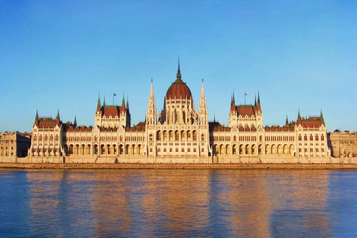 Hungarian Parliament Building with red-tiled dome and Gothic spires reflected in the Danube River against blue sky
