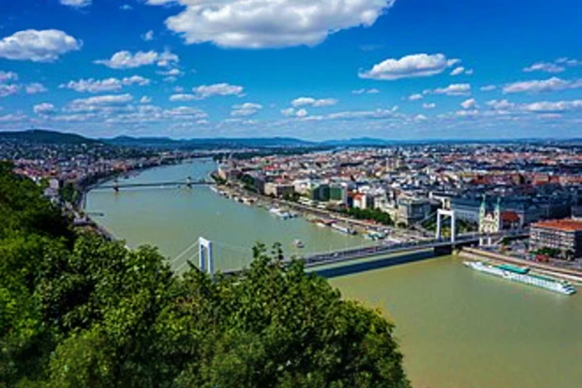 Aerial view of Budapest with Danube River, suspension bridge, city skyline, and forested hillside under blue sky with white c