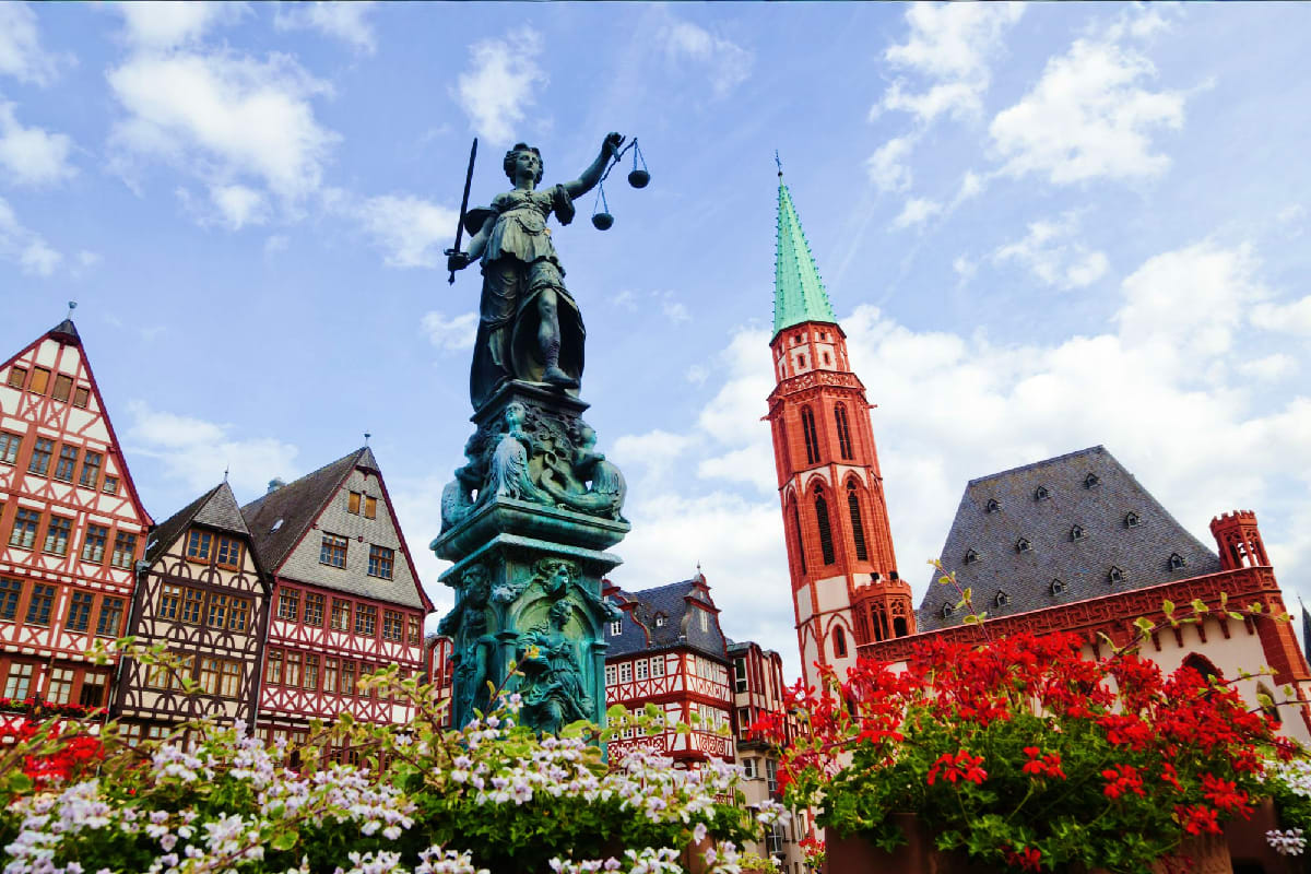 Justice statue in Frankfurt's Römerberg square with historic half-timbered buildings, red church tower, and flowering gardens