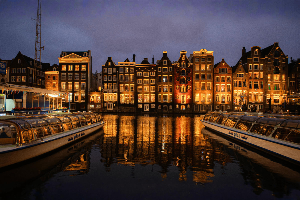 Historic Amsterdam buildings lit up at dusk reflecting in canal water with tour boats moored in foreground