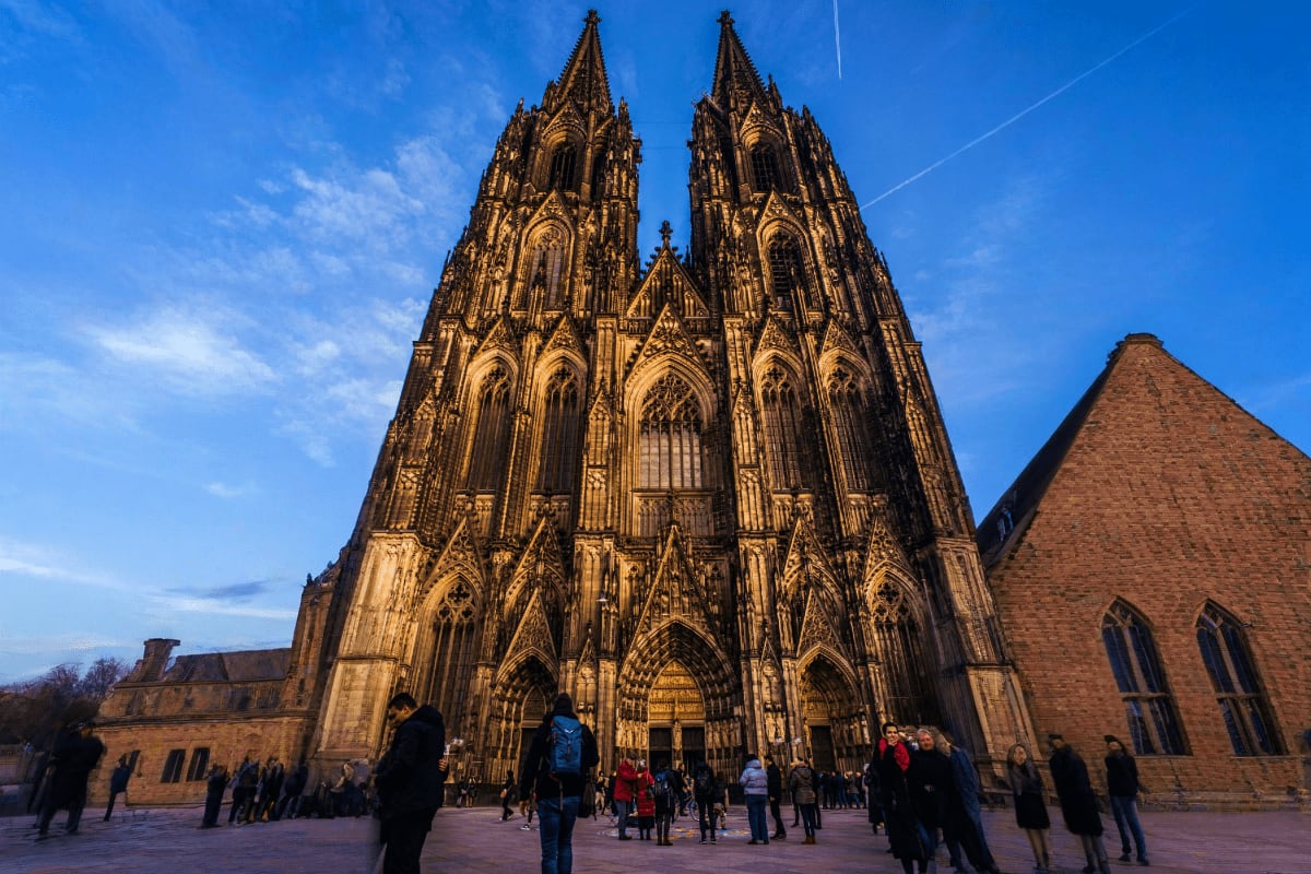 Cologne Cathedral Gothic twin spires with ornate stone details against blue sky, tourists gathering in plaza below