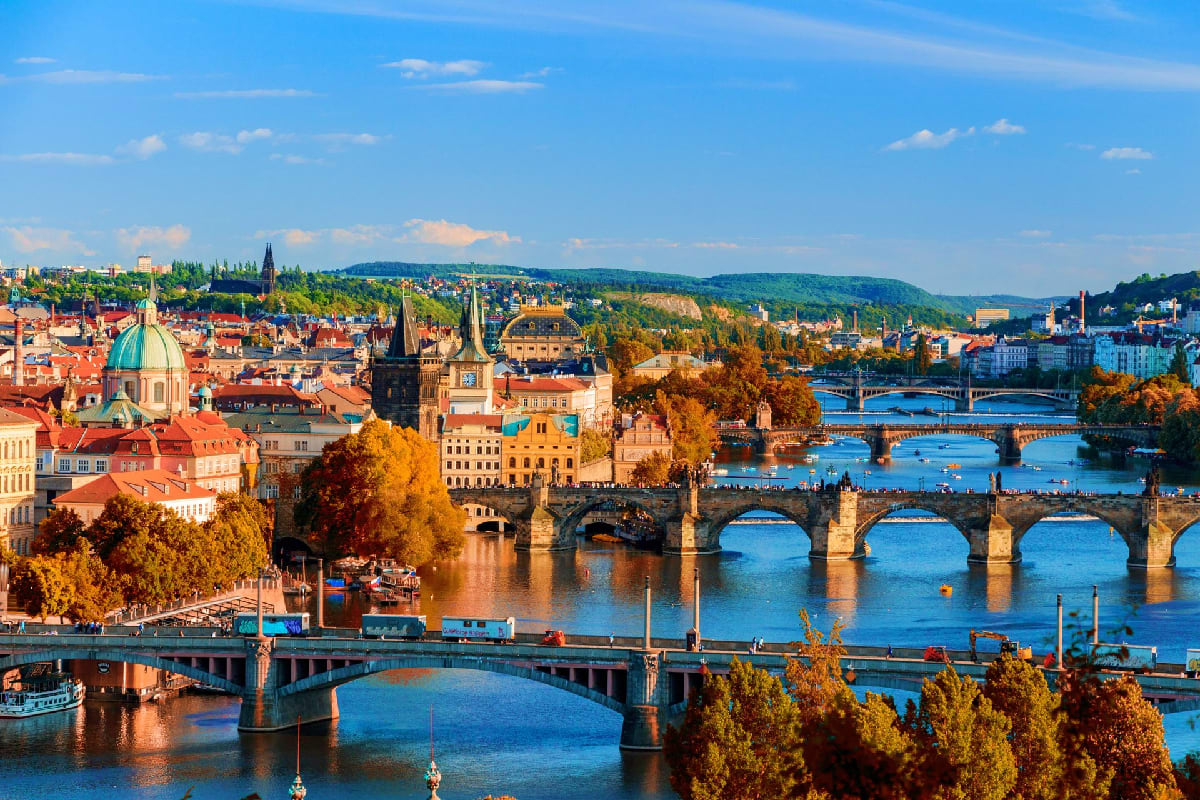 Prague cityscape with Charles Bridge spanning the Vltava River, historic red-roofed buildings, church spires, and autumn foli
