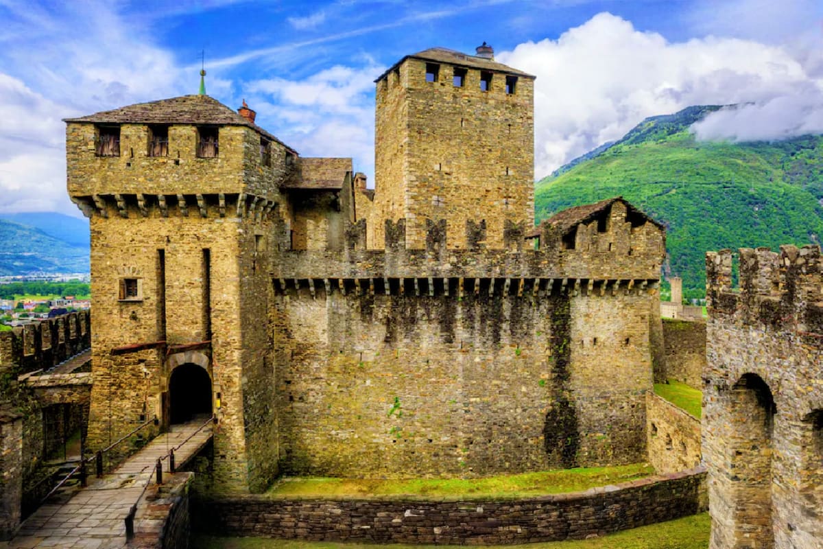 Medieval stone castle with towers and battlements overlooking green Alpine mountains and valley landscape under blue sky