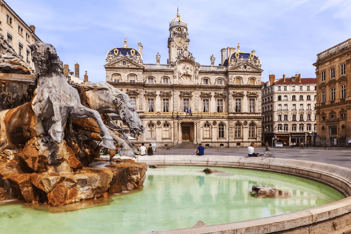 Ornate stone fountain with sculptural figures in foreground, historic Beaux-Arts building with clock tower and golden dome in