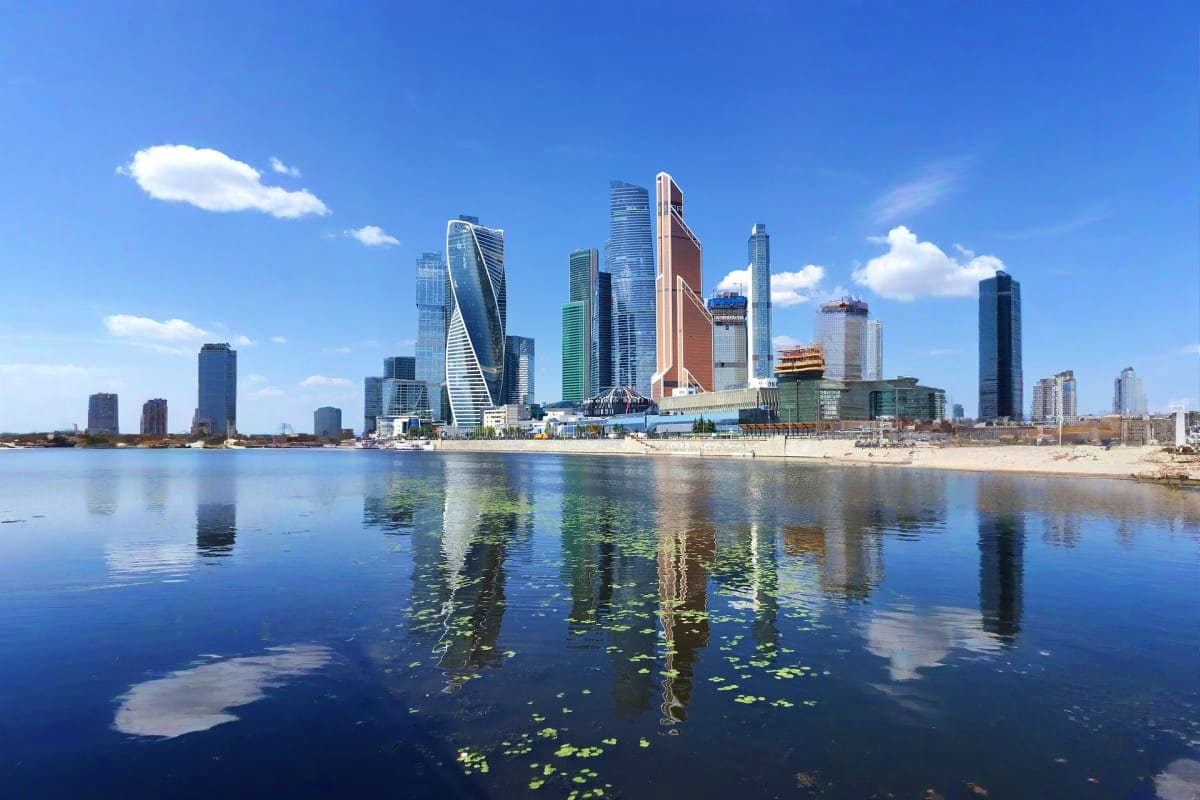 Modern city skyline with high-rise buildings reflected in calm blue water under clear sky with white clouds