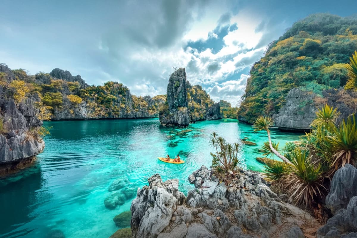 Turquoise lagoon surrounded by limestone cliffs and palm trees with kayakers exploring the crystal-clear water in El Nido, Ph