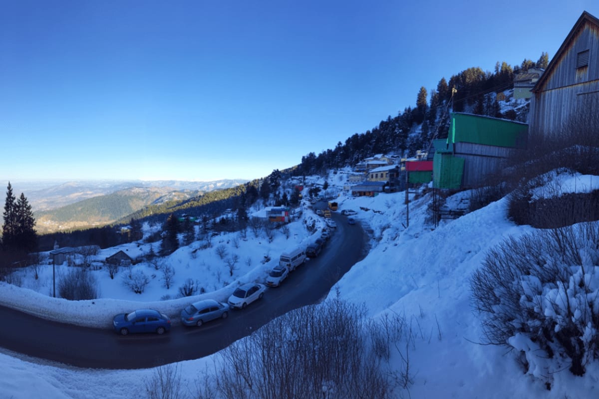 Snow-covered mountain village with parked cars on winding road, forested slopes, and valley view below in winter conditions