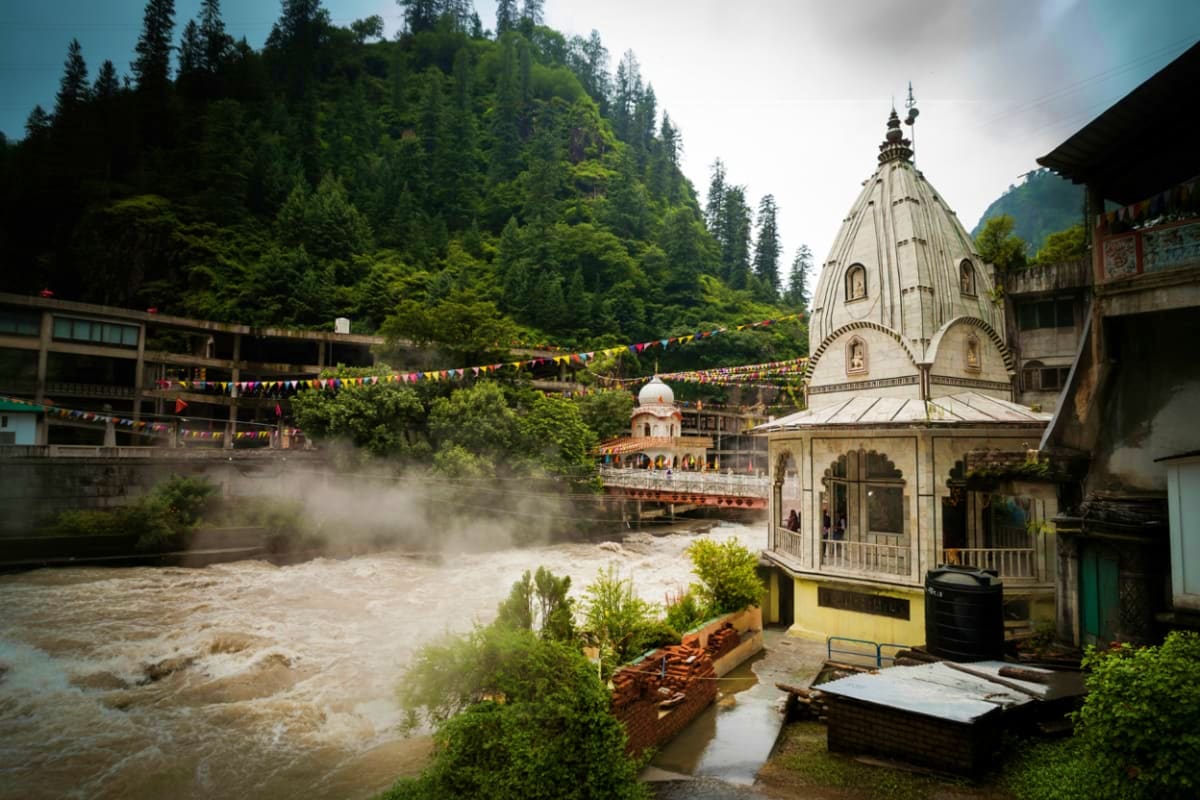 Hindu temple with white dome beside rushing river in mountainous forest valley with colorful prayer flags and traditional bui