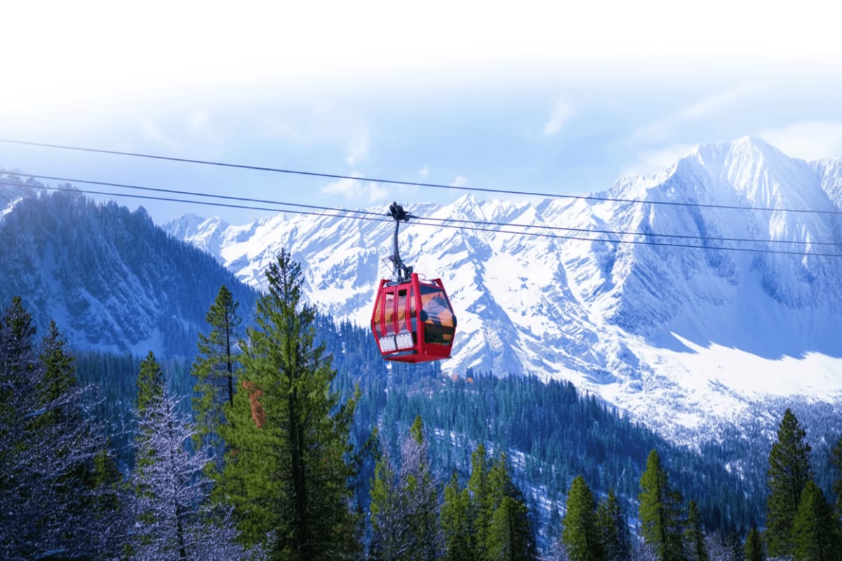 Red cable car suspended above snow-covered mountains and evergreen forests in a mountain valley landscape.