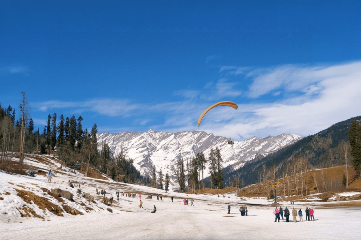 Paraglider soaring over snowy Solang Valley with snow-capped Himalayan peaks, forested slopes, and tourists enjoying winter a