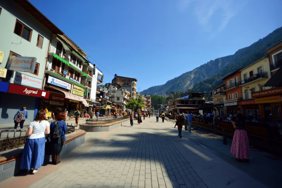 Bustling mountain town street in Manali with colorful buildings, shops, and tourists under clear blue sky with forested mount