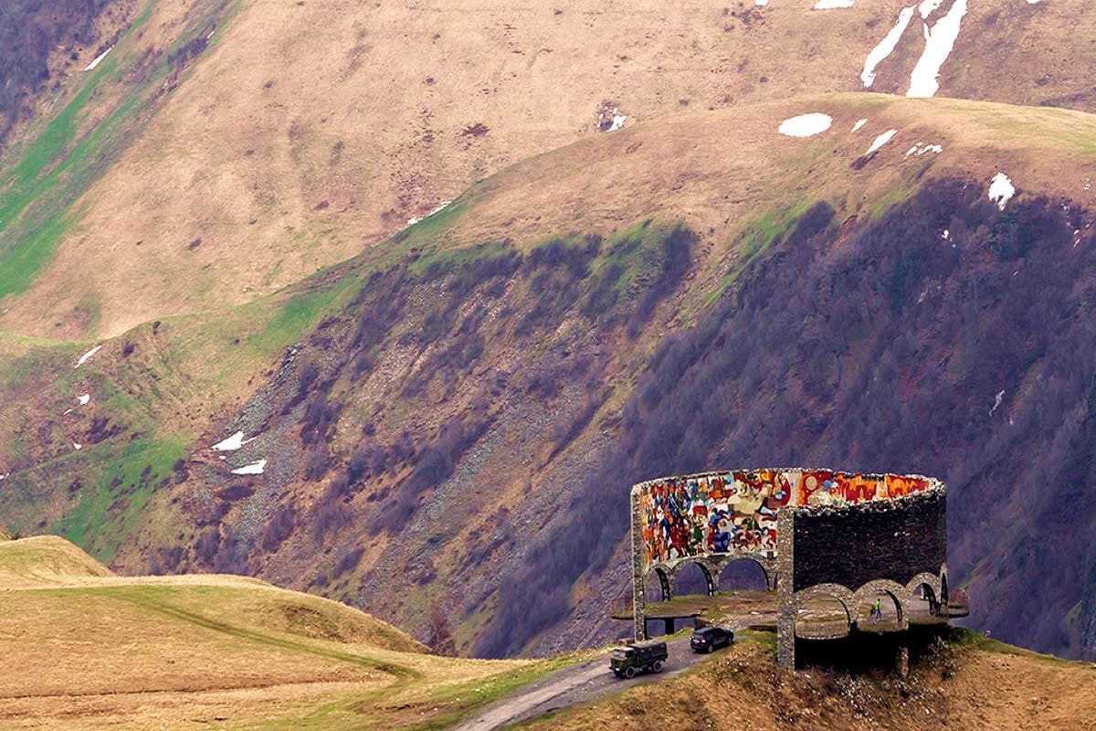 Abandoned Soviet-era structure with colorful murals on mountainside in Georgian highlands with snow patches and winding road