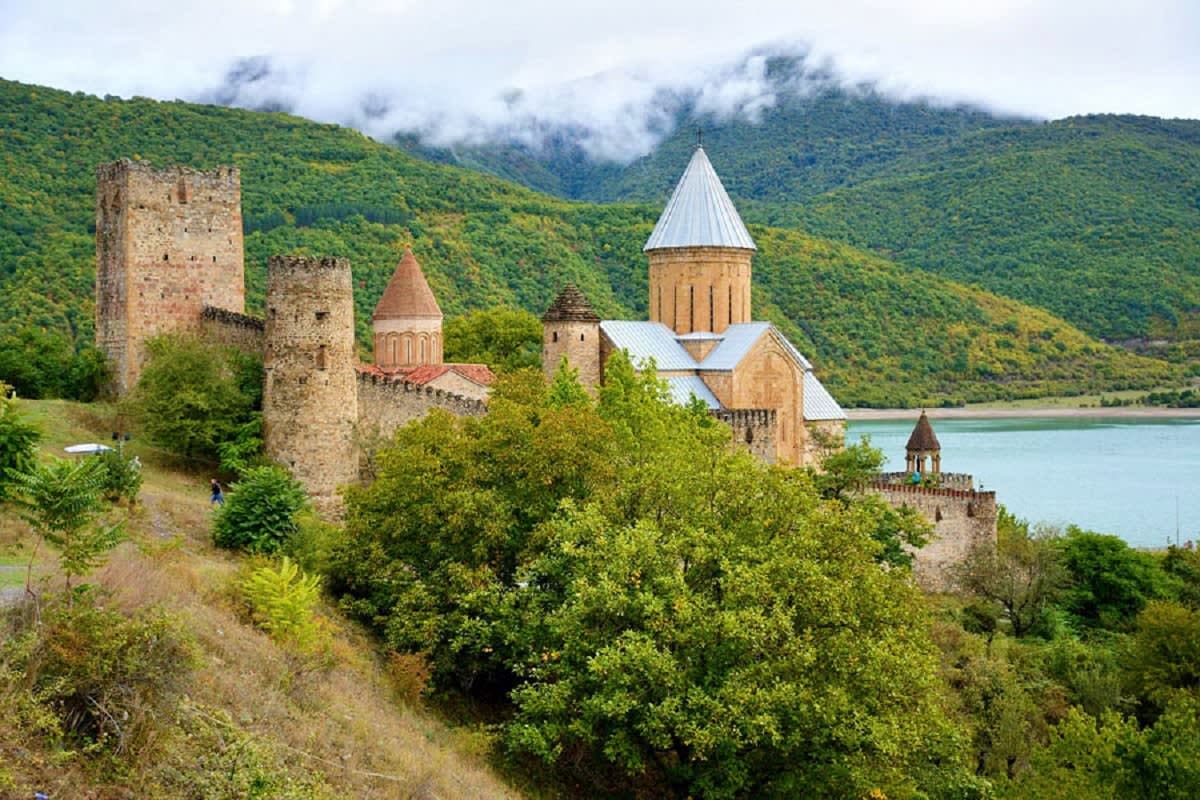 Medieval fortress and church with cone-shaped roofs overlooking a turquoise lake surrounded by green mountains in Georgia