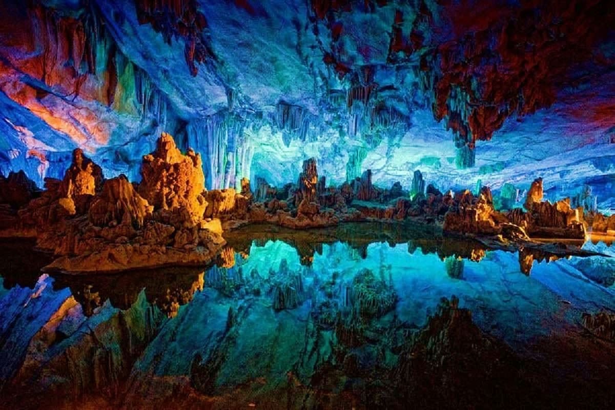 Underground cave with turquoise pool reflecting blue-lit stalactites and stalagmites in golden-orange stone formations