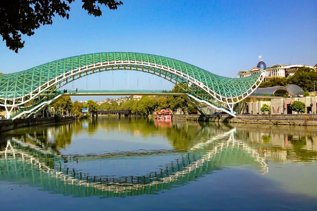 Green and white arched bridge reflected in calm river water with historic buildings and trees in Tbilisi, Georgia