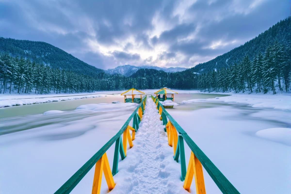 Wooden bridge with green and yellow railings crossing frozen lake surrounded by snow-covered pine forests and mountains in wi