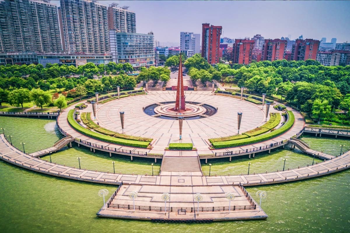 Aerial view of a circular waterfront plaza with red monument in center, surrounded by green landscaping and modern high-rise 