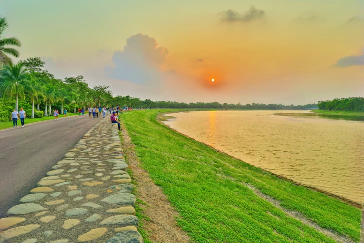 Riverbank promenade at sunset with palm trees, green lawn, stone pathway, and visitors walking along the water's edge
