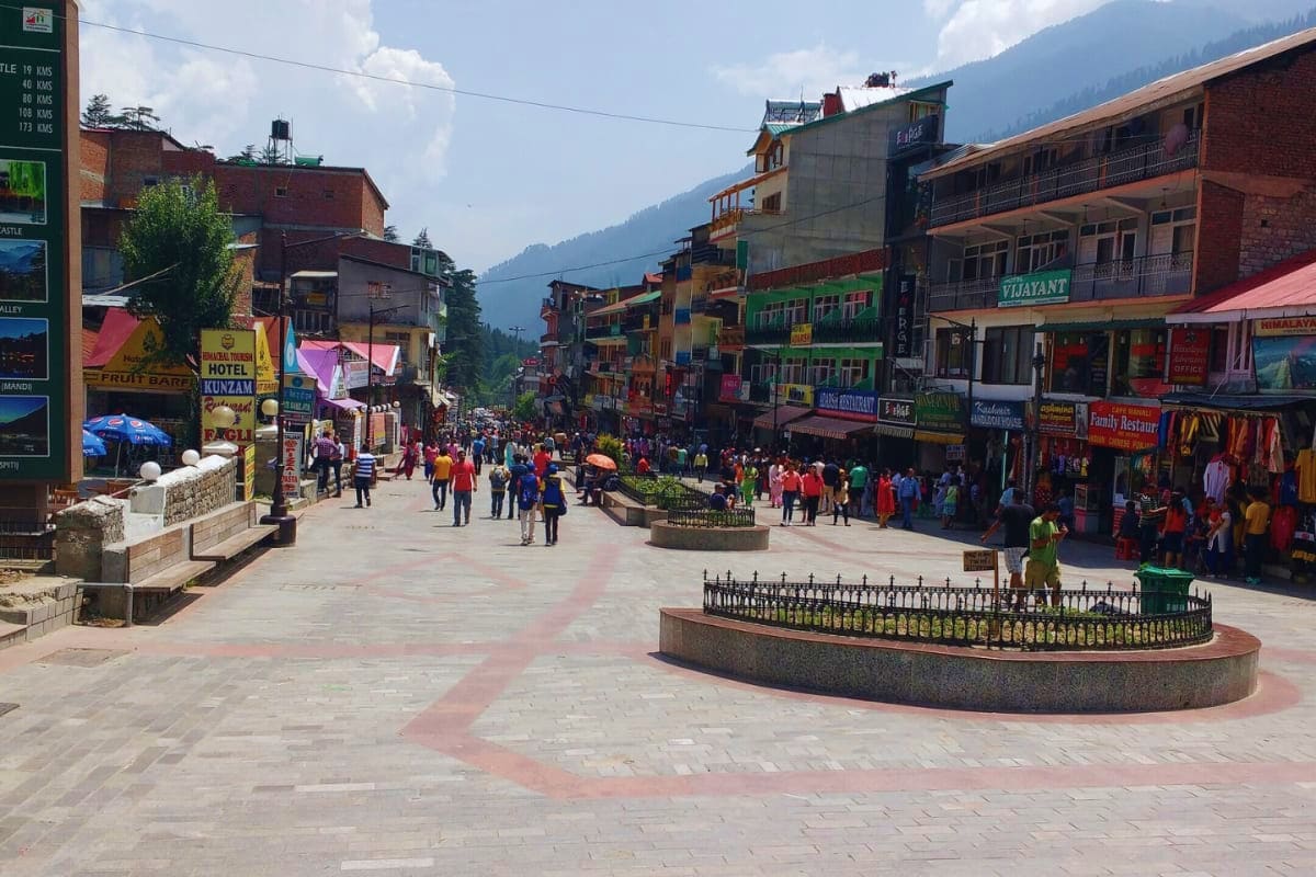 Busy mountain town square in Himachal Pradesh with colorful buildings, shops, tourists, and forested hills in the background