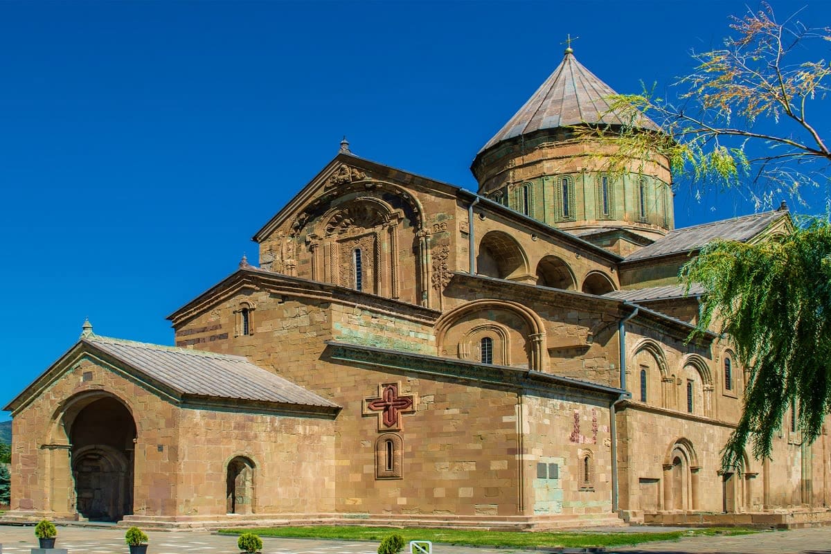 Georgian Wellness Escape Historic stone church with conical dome and cross, Georgian architecture under blue sky with green foliage