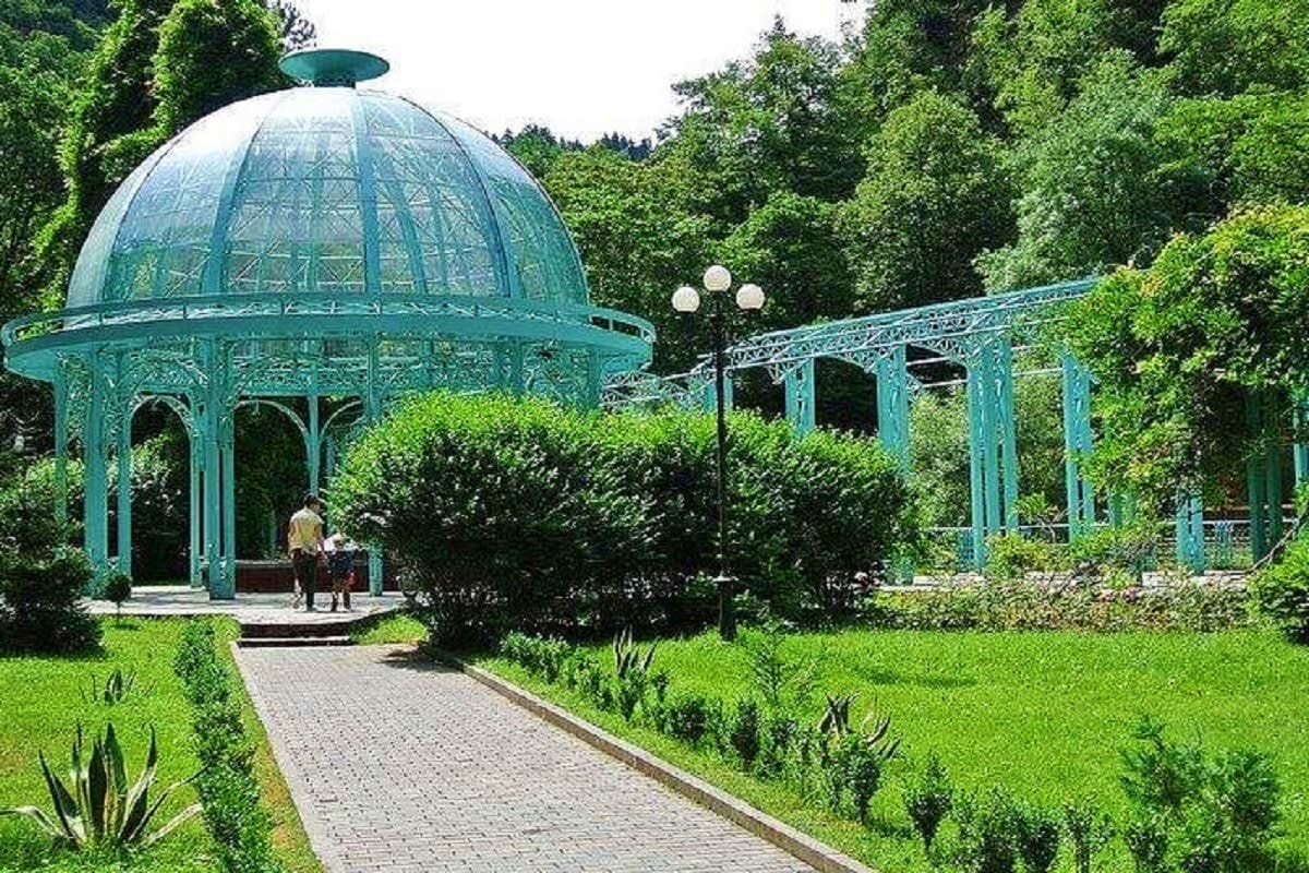 Georgian Wellness Escape Turquoise glass dome and pavilion structures in a lush green garden with walking paths and hedges, surrounded by forest trees