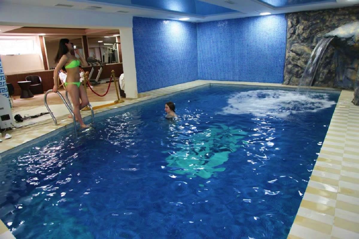 Indoor swimming pool with blue tile walls, woman swimming, another woman standing on pool deck in green bikini, waterfall mur