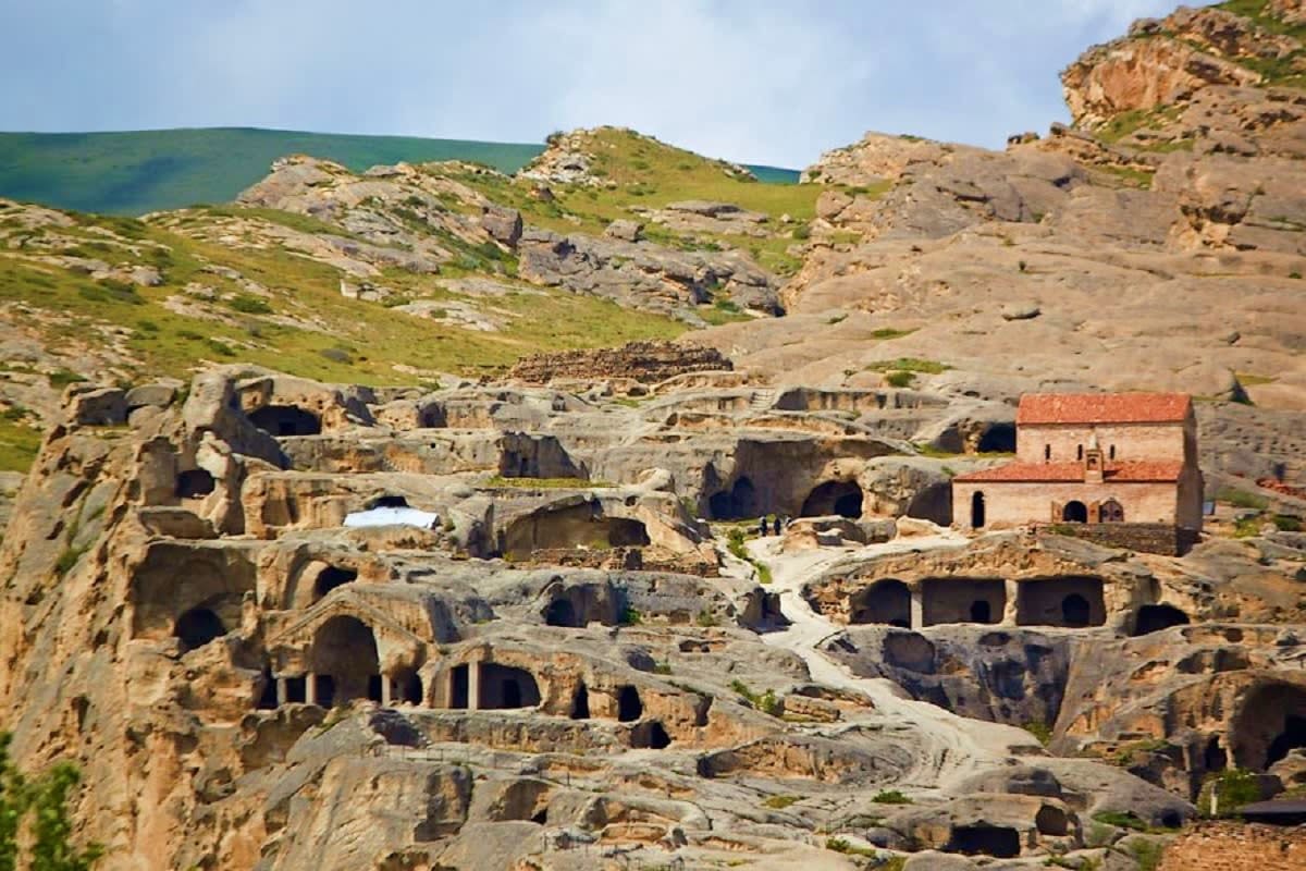 Ancient cave dwellings carved into limestone cliffs with a red-brick church building nestled among rocky terrain and green hi