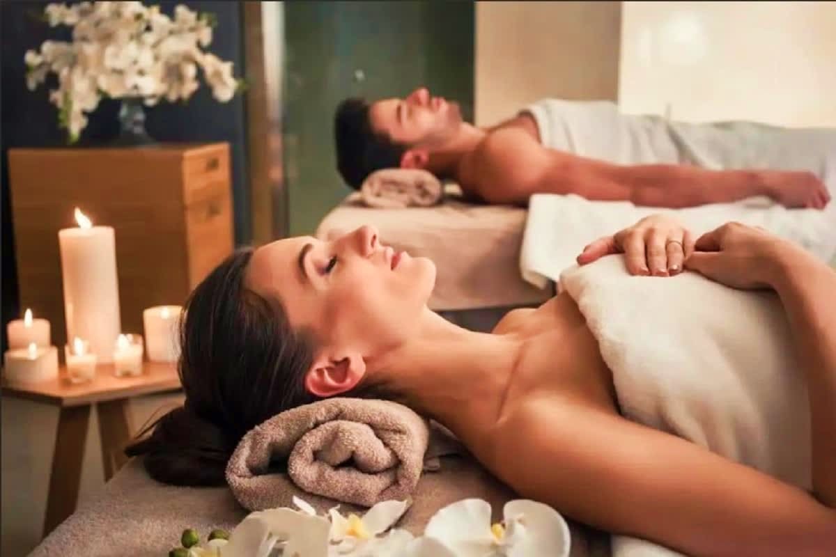 Two people relaxing on massage tables in a spa setting with lit candles, rolled towels, and white orchid flowers