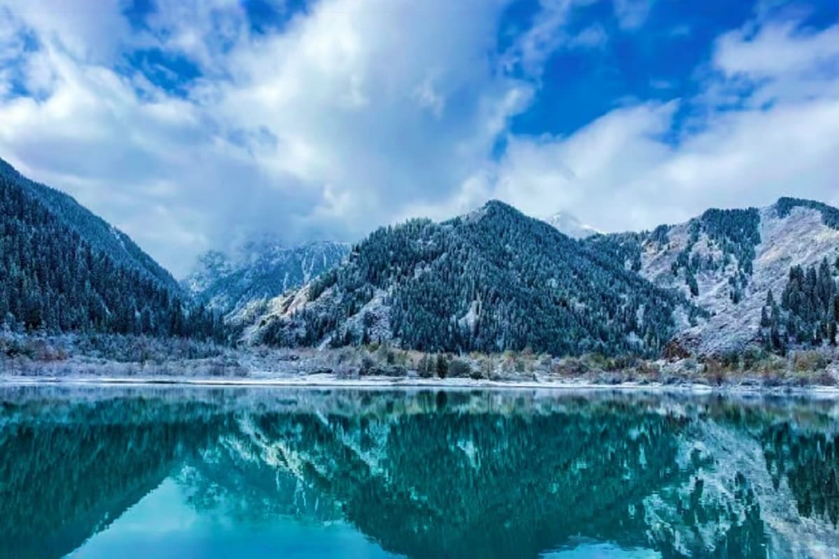 Mountain lake with coniferous forest and snow-capped peaks reflecting in turquoise water under blue sky with clouds